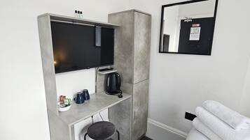 Quarto de hotel compacto com cama, escrivaninha com televisão, um banquinho, lixeira e espelho.