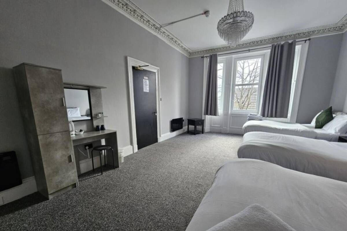 A hotel room with a large bed, a desk with a chair, a wardrobe, a mirror, a chandelier, and a window with curtains.