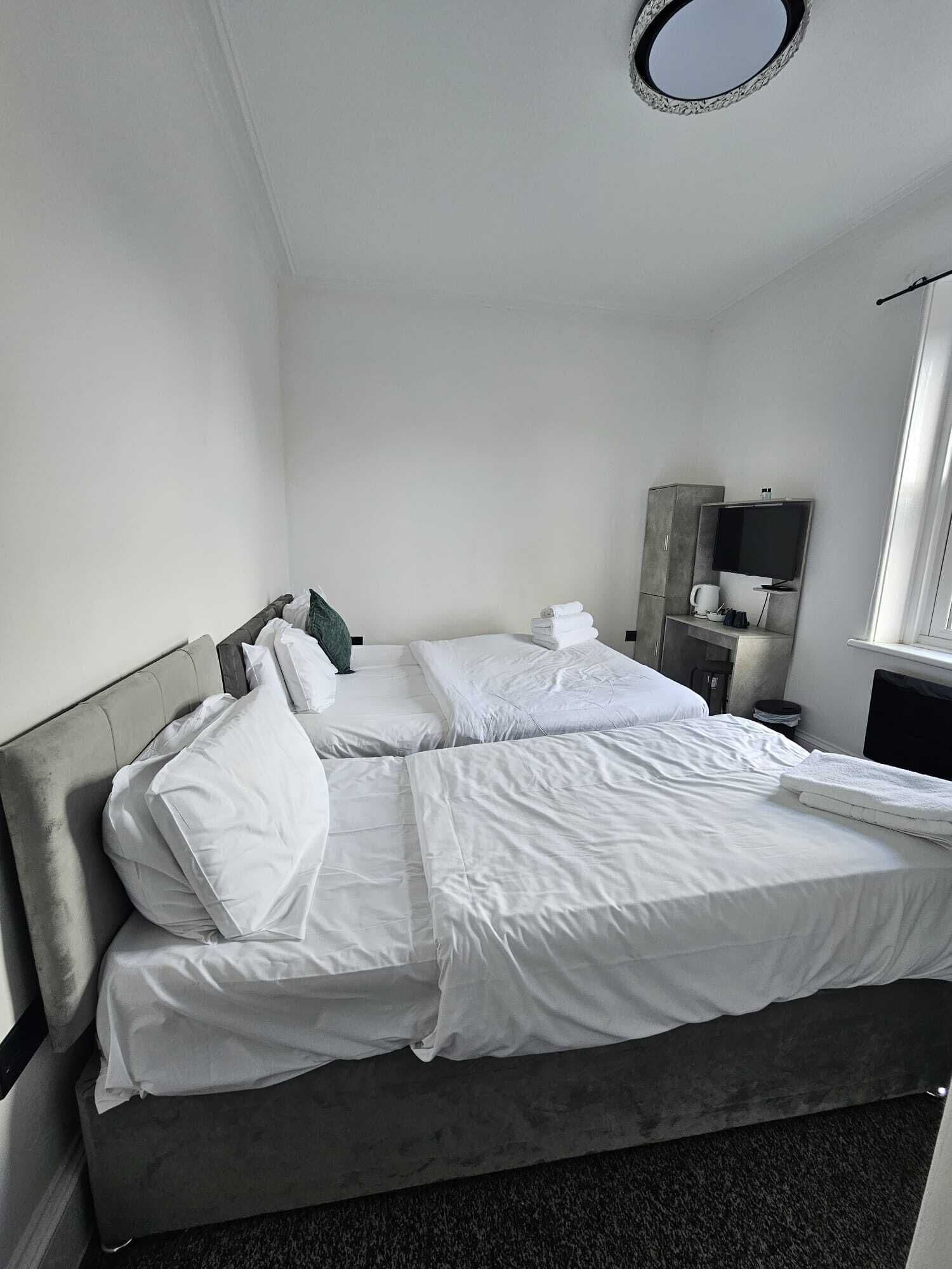 A single bed with white linens, a grey headboard, a bedside table with a lamp, and a television on a stand.