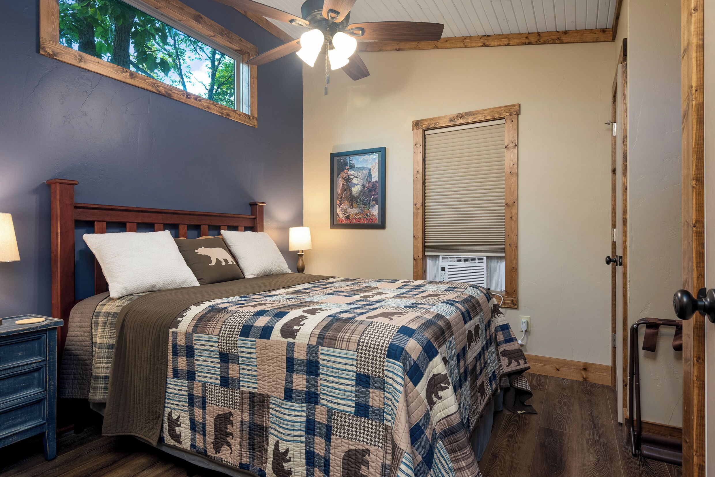 A bedroom with a wooden bed, a ceiling fan, a window with a view of trees, and a door with a wooden frame.