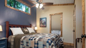 A bedroom with a wooden bed, a ceiling fan, a window with a view of trees, and a door with a wooden frame. - Cozy Ozark Cottage Near Spring River & Mammoth Spring (Mammoth Spring)