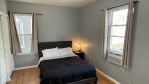 Een slaapkamer met een bed, een nachtkastje met een lamp, een raam met gordijnen en een wandairconditioner.