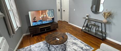 Een woonkamer met een flatscreen televisie, een salontafel, een bank, een console tafel met spiegel, en een houten vloer.