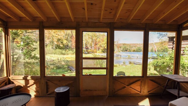 Ein Interieur einer Holzhütte mit großen Fenstern, Holzbalken und Blick auf einen See und einen Wald.