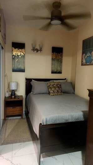 Ein Schlafzimmer mit einem Bett, einem Nachttisch, einer Lampe, einem Deckenventilator und Kunstwerken an den Wänden.
