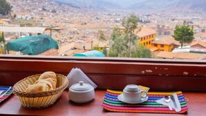 A window view of a cityscape with a table featuring a basket of bread, a teapot, and a colorful mat. - Mirador All Sur Cusco - Santa Ana (Cusco)