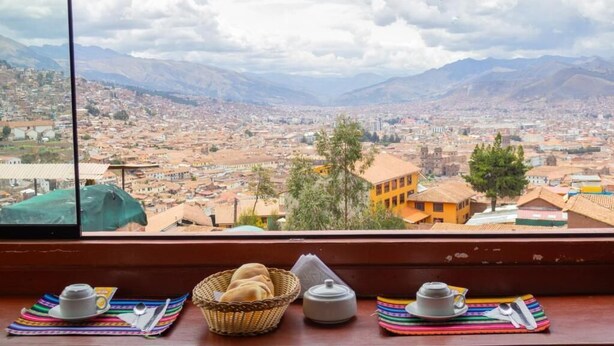 A cityscape with mountains in the background, viewed from a window with a table featuring a basket of bread, a teapot, and a cup. - Mirador All Sur Cusco - Santa Ana (Cusco)