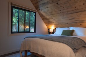 Een slaapkamer met een houten plafond, een groot raam, een bed met kussens en een bedlampje.