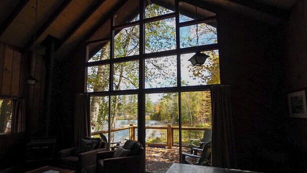 A cabin interior with large windows, wooden beams, a fireplace, and a view of a lake and trees. - Lakeside Adirondack Resort-Cabin 7A (Olmstedville)
