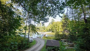 A forested area with a dirt path leading to a small building and a bridge over a body of water. - Lakeside Adirondack Resort-Cabin 7A (Olmstedville)