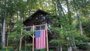 A wooden cabin surrounded by trees with an American flag hanging in front. - Lakeside Adirondack Resort-Cabin 7A (Olmstedville)