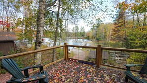A wooden deck with chairs, a railing, and a view of a lake and forest. - Lakeside Adirondack Resort-Cabin 7A (Olmstedville)