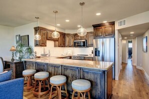 A kitchen with wooden cabinets, a central island with stools, pendant lights, and stainless steel appliances. - Sea Gull #202LK: Beachfront 1 Bedroom 1 Bathroom With Spectacular Views and 24 Hour Management (Port Aransas)