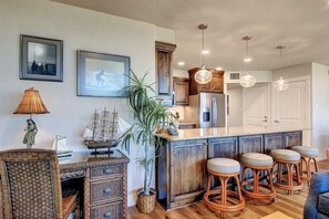 A kitchen with wooden cabinets, a central island with stools, pendant lights, and framed pictures on the wall. - Sea Gull #202LK: Beachfront 1 Bedroom 1 Bathroom With Spectacular Views and 24 Hour Management (Port Aransas)