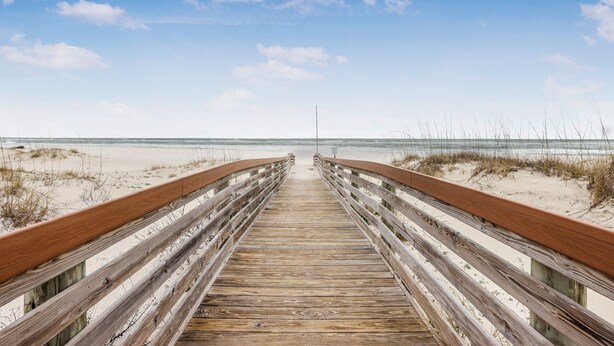 Een houten looppad dat naar een strand met duinen en een heldere lucht leidt.