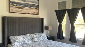 A bedroom with a bed, a ceiling fan, a painting, a nightstand, a lamp, and a window with curtains. - New Rental ~ Spacious Beach House - Hot Tub ~ Walk to the Beach (Kitty Hawk)