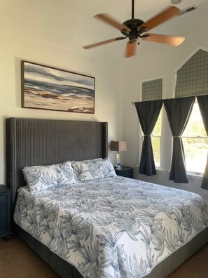 A bedroom with a bed, a ceiling fan, a painting, a nightstand, a lamp, and a window with curtains. - New Rental ~ Spacious Beach House - Hot Tub ~ Walk to the Beach (Kitty Hawk)