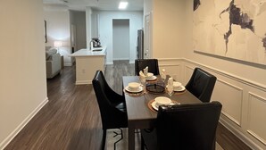 Un espace repas avec une table rectangulaire dressée pour quatre personnes, des chaises noires, un tapis à motifs géométriques et une œuvre d’art abstraite accrochée au mur.