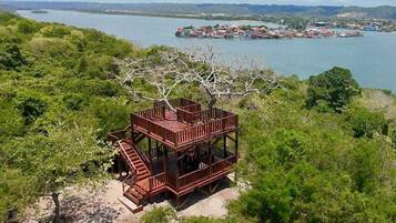 A multi-level wooden structure with a staircase, surrounded by dense greenery and overlooking a body of water with a distant settlement.