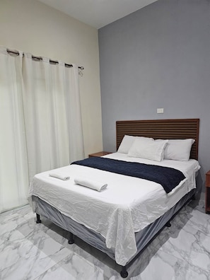 A neatly made bed with white bedding and a dark blue throw, a wooden headboard, and a marble-patterned floor.