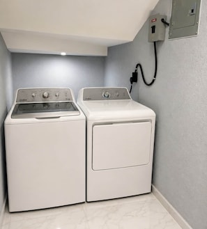 A laundry room with two white washing machines, a control panel, and a power outlet on the wall.