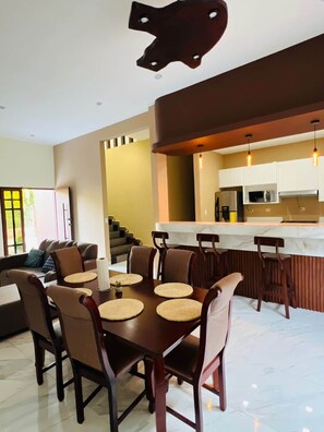 A modern kitchen with a central island, dining area with a table and chairs, and a living room with a sofa and a staircase.