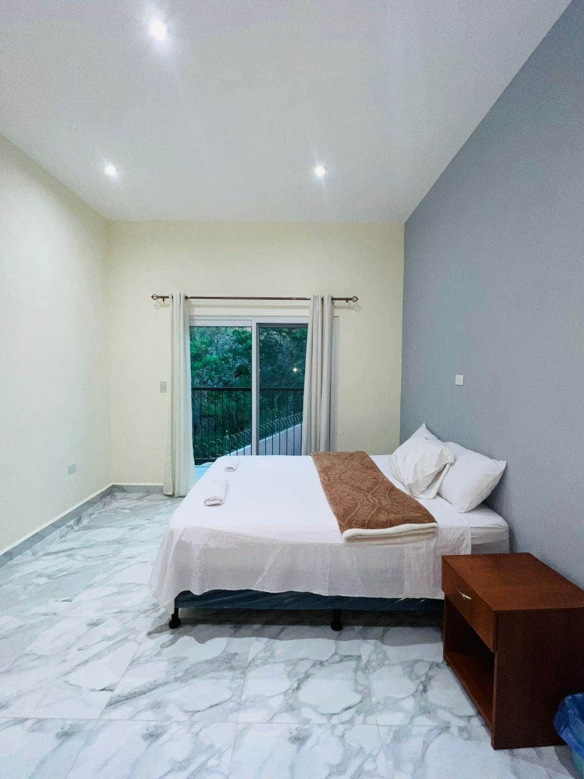 A bedroom with a bed, a nightstand, a window with curtains, and a view of greenery outside.