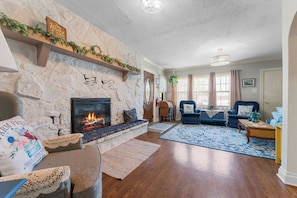 A living room with a stone fireplace, wooden flooring, a blue patterned rug, and a blue armchair.