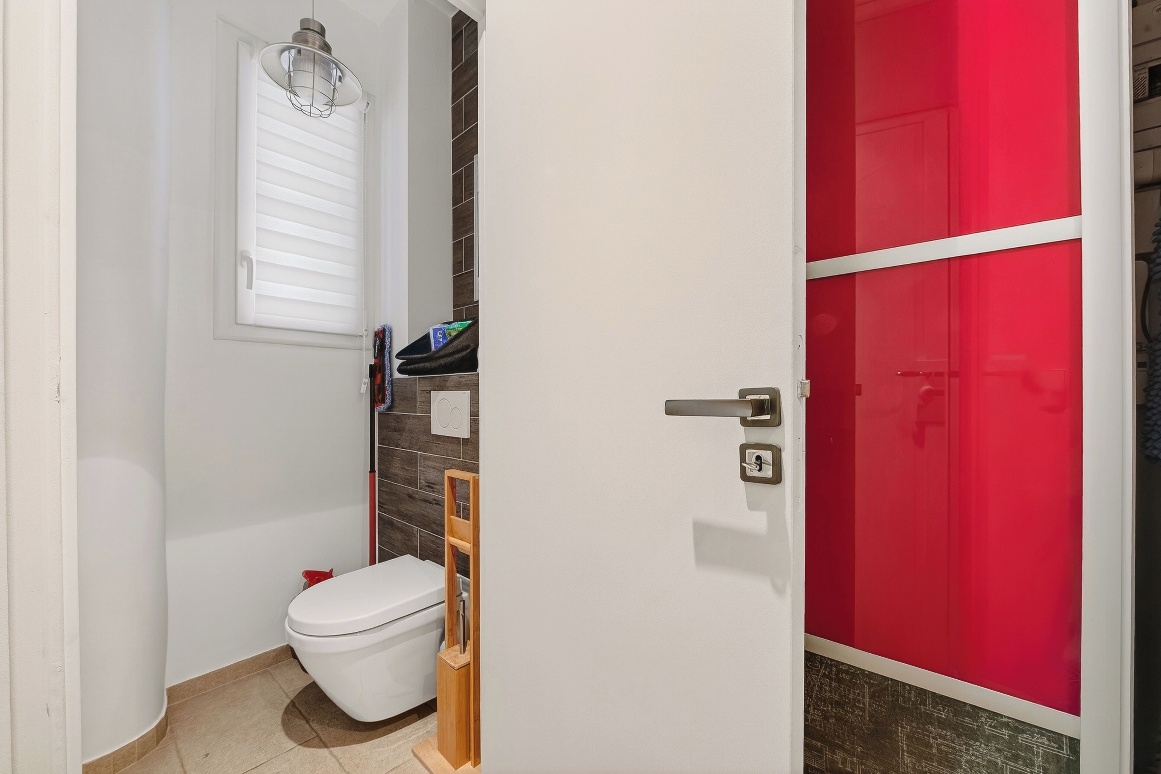 A bathroom with a toilet, a shelf with toiletries, a window with blinds, and a red door.
