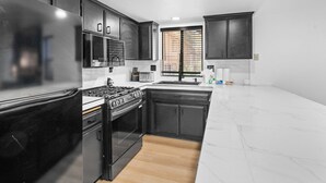 Una cocina con armarios oscuros, un refrigerador de acero inoxidable, una estufa y una ventana con persianas.