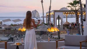 A woman in a white dress holding a glass of wine, overlooking a beach with lounge chairs and a fire feature. - Cabo San Lucas oceanfront getaway with resort amenities (Cabo San Lucas)