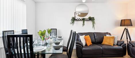 Una sala moderna con una mesa de comedor de vidrio, sillas negras, un sofá de cuero, un tapete estampado, un espejo y una lámpara de pie.