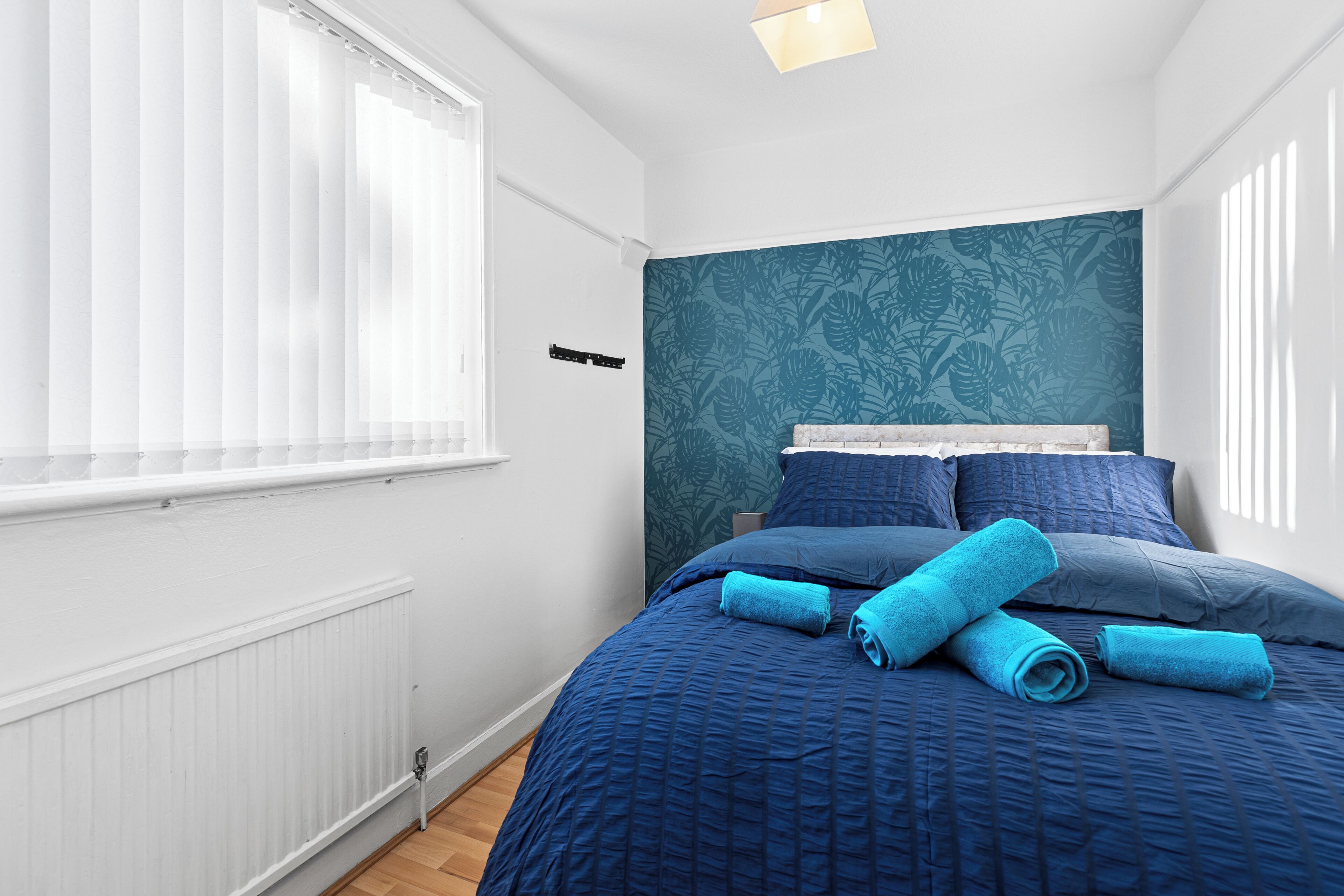 Een slaapkamer met een bed, blauw beddengoed, een radiator en een raam met zonwering.