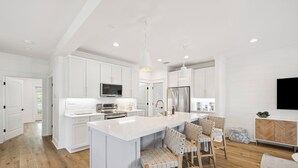 A modern kitchen with white cabinetry, a central island, wooden chairs, and a built-in TV. - 🎥 Outdoor Movie Screen | Fire Pit | Games | Bikes | Sleeps 10 - Bike to Beach! (Inlet Beach)