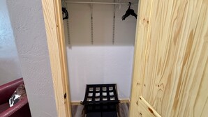 A wooden closet with a bench, hanging clothes, and a metal shelf.