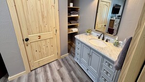A bathroom with a wooden door, a vanity with a sink, a mirror, and shelves with towels.