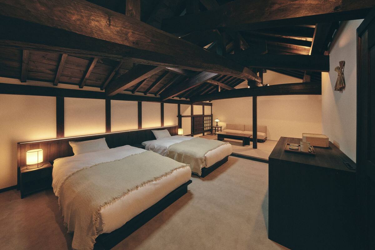 A room with two beds, a nightstand with a lamp, a small table, and a chair.