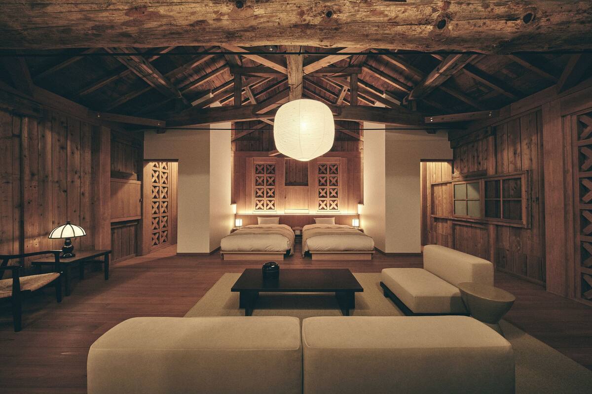 A room with wooden walls, a high ceiling, a large hanging light fixture, two beds, a sofa, a coffee table, and a small table with a lamp.