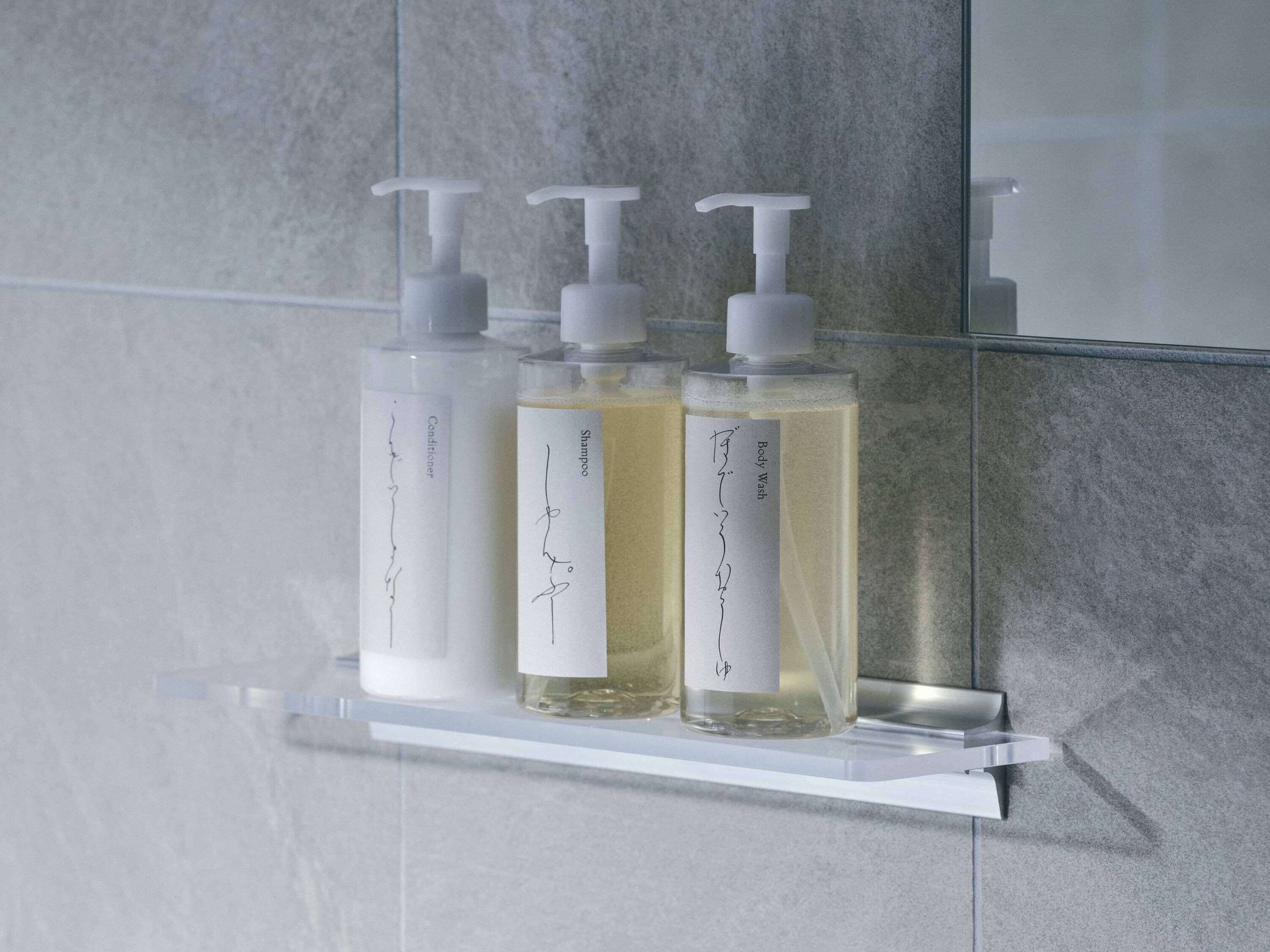 Three bottles of liquid on a clear shelf against a tiled wall.