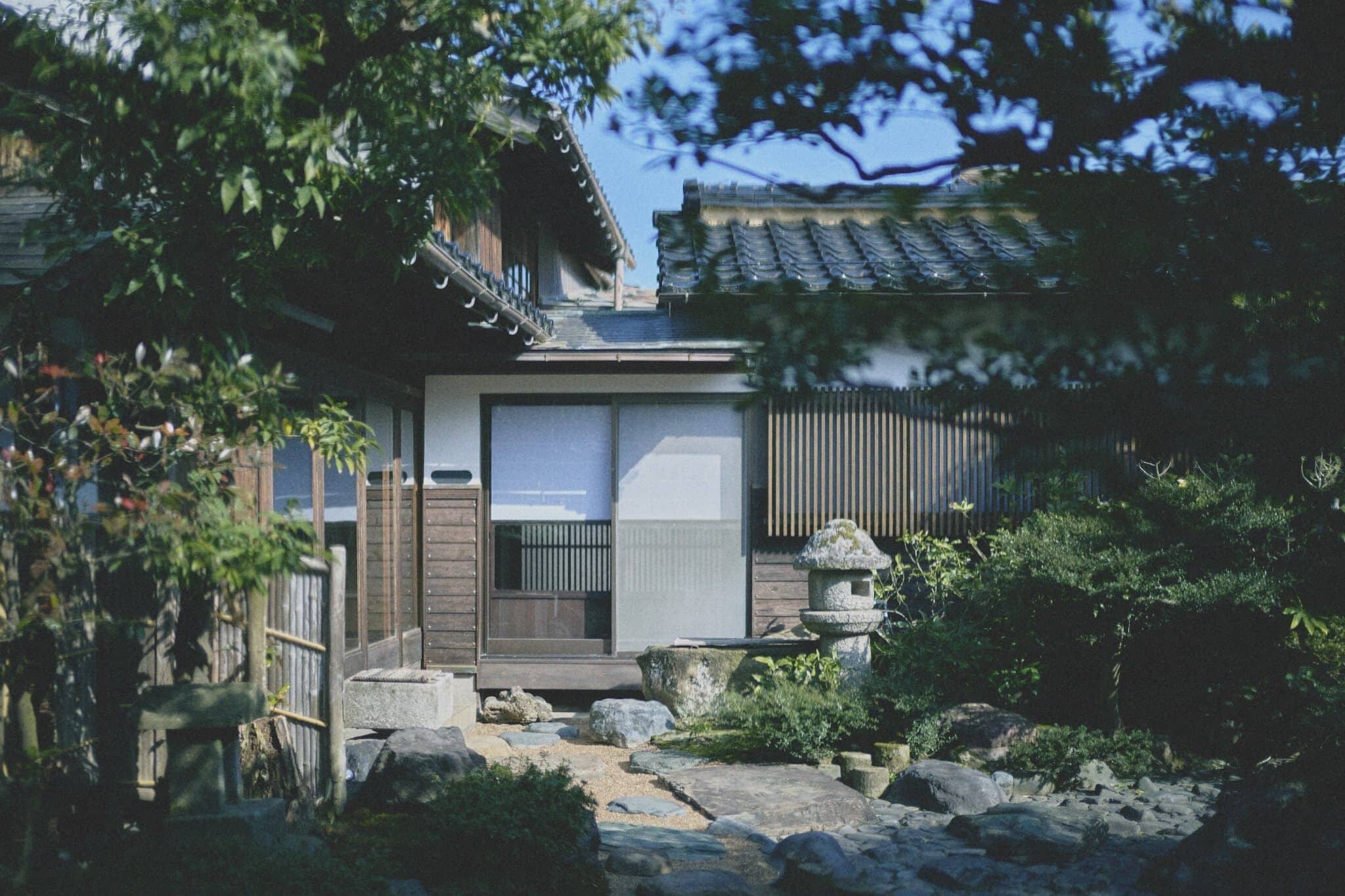 Una casa tradicional japonesa con fachada de madera, un farol de piedra y un jardín con rocas y plantas.