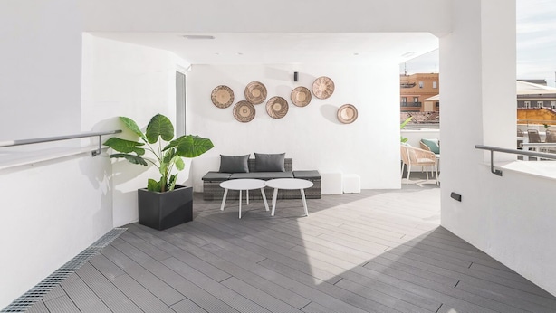 Een modern balkon met buitengebeuren, een grijze houten vloer, een grijze bank, twee witte ronde tafels en een grote pot met planten.