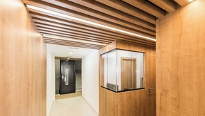 Een hal met houten wanden en plafond, een glazen lift en een deur naar een andere kamer.