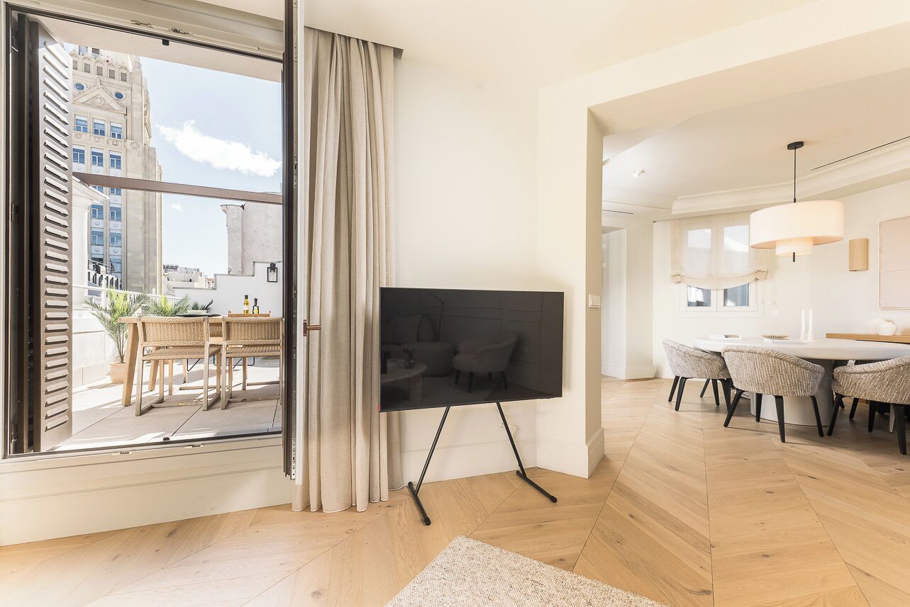 Een moderne woonkamer met een groot raam, een flatscreen televisie op een standaard, een zithoek met rieten stoelen en uitzicht op een stadsbeeld.