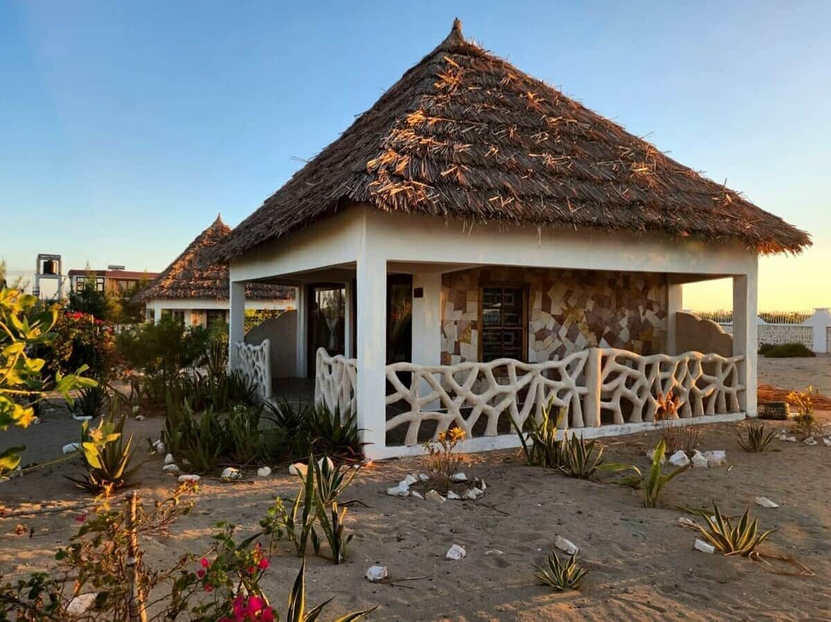 Una pequeña estructura de techo de paja con exterior blanco y una cerca decorativa, rodeada de vegetación desértica.