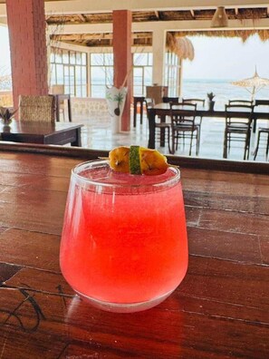 A glass of pink drink with a garnish on a wooden table in a room with a view of the ocean.