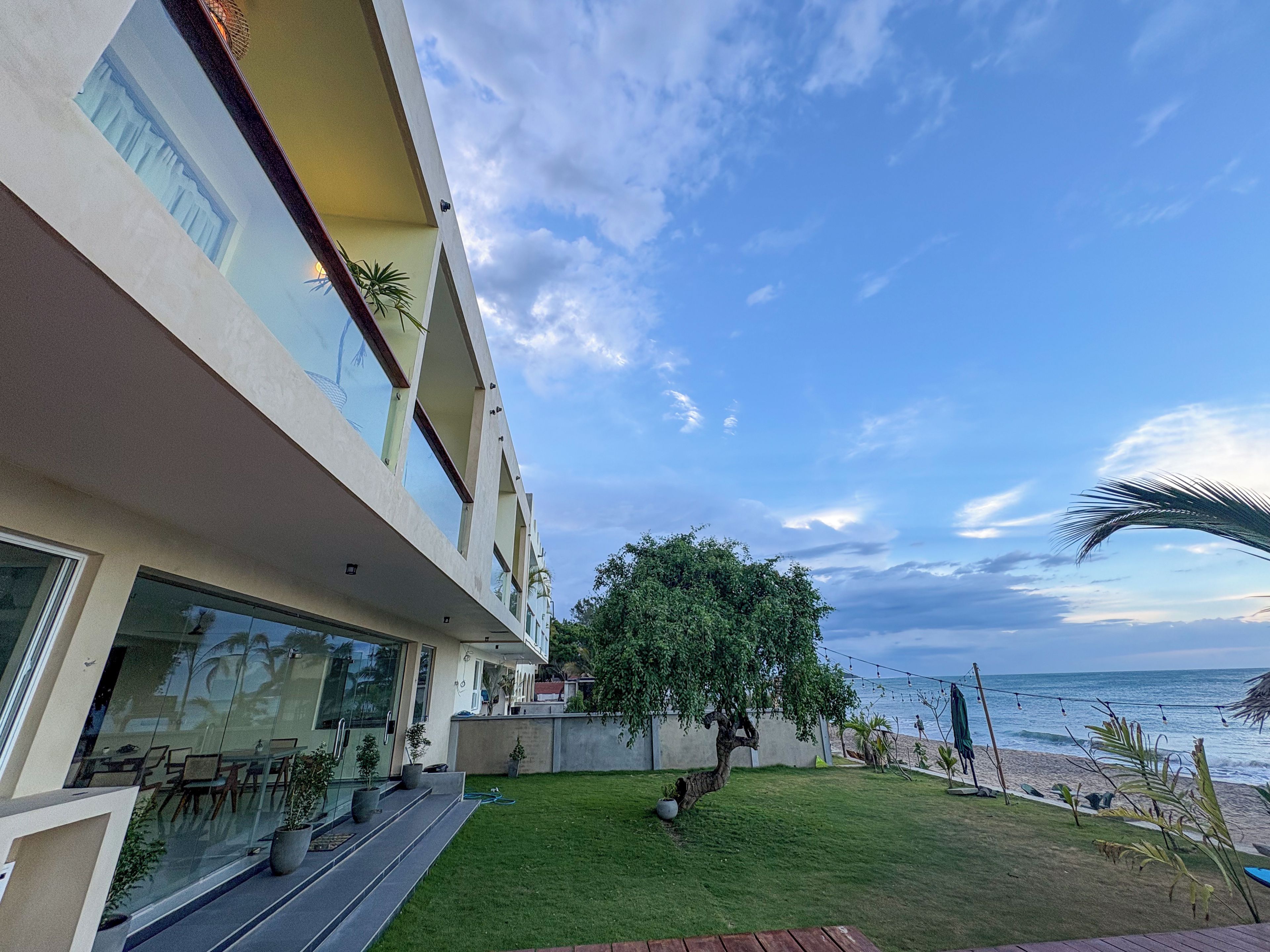 A modern building with a flat roof, large windows, and a balcony overlooking a grassy area with a tree and a view of the sea.