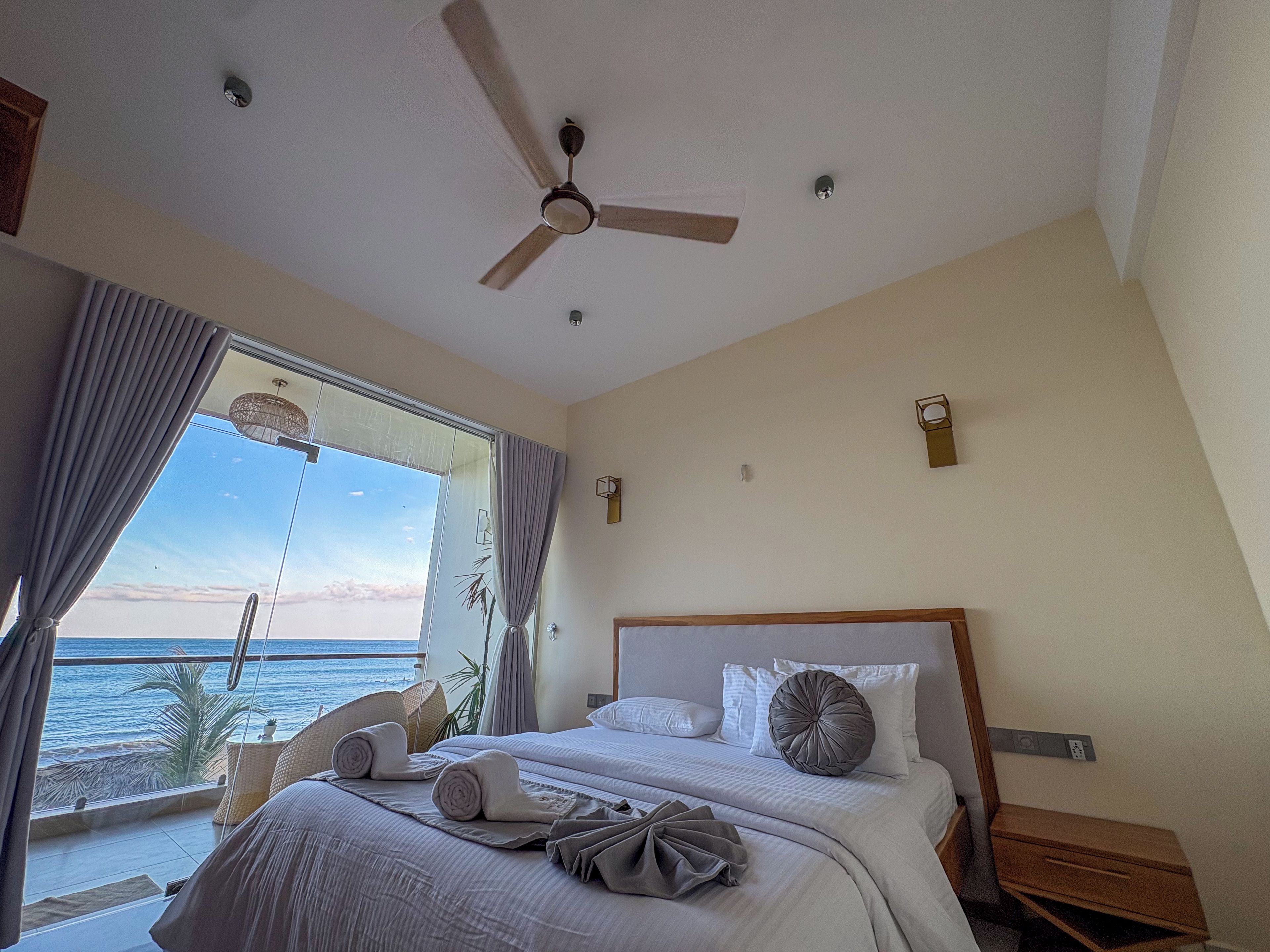 Habitación de hotel con cama, ventilador de techo, vista al mar y ventana con cortinas.
