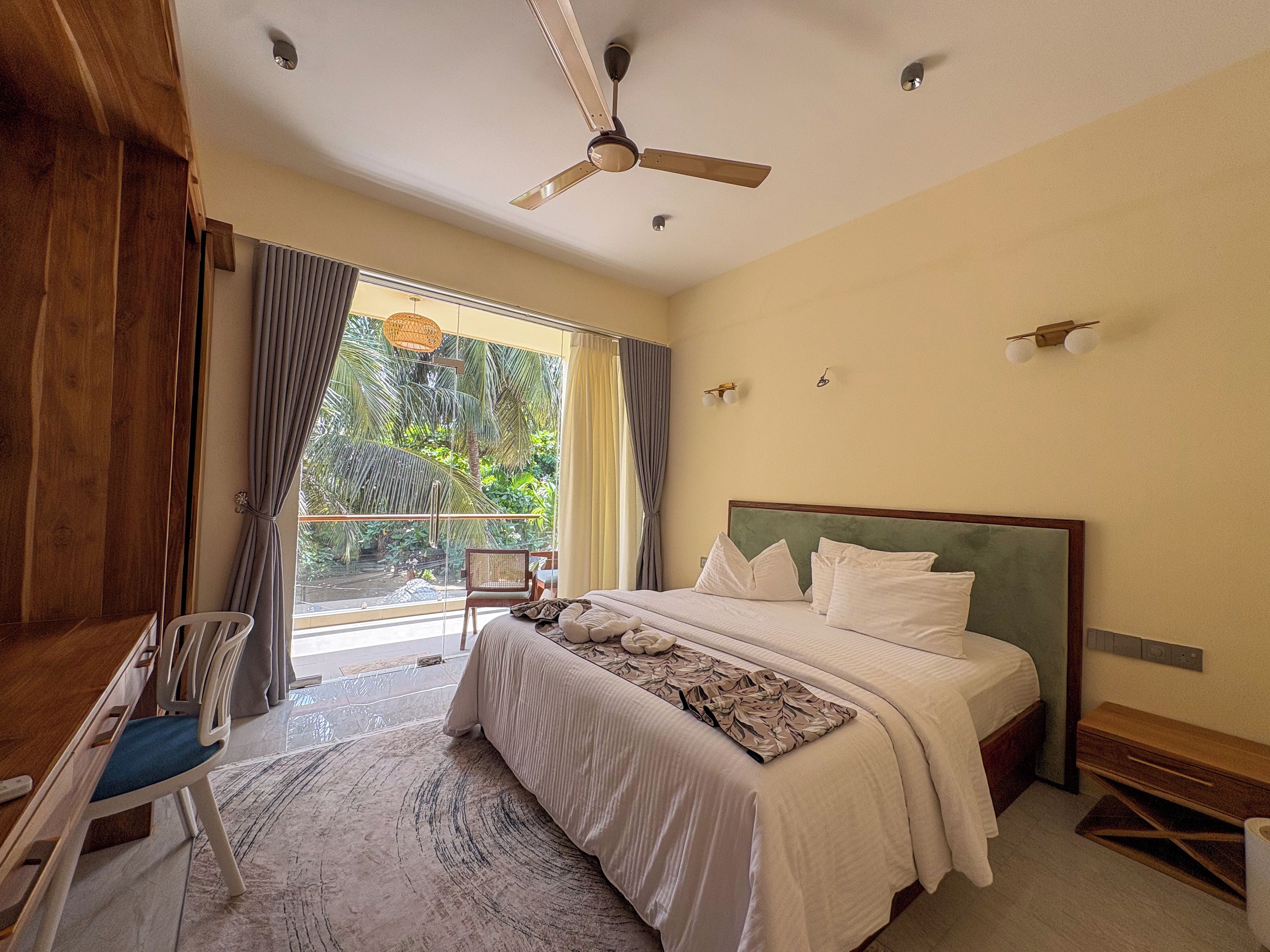 A hotel room with a bed, a desk, a chair, a ceiling fan, and a view of greenery outside.