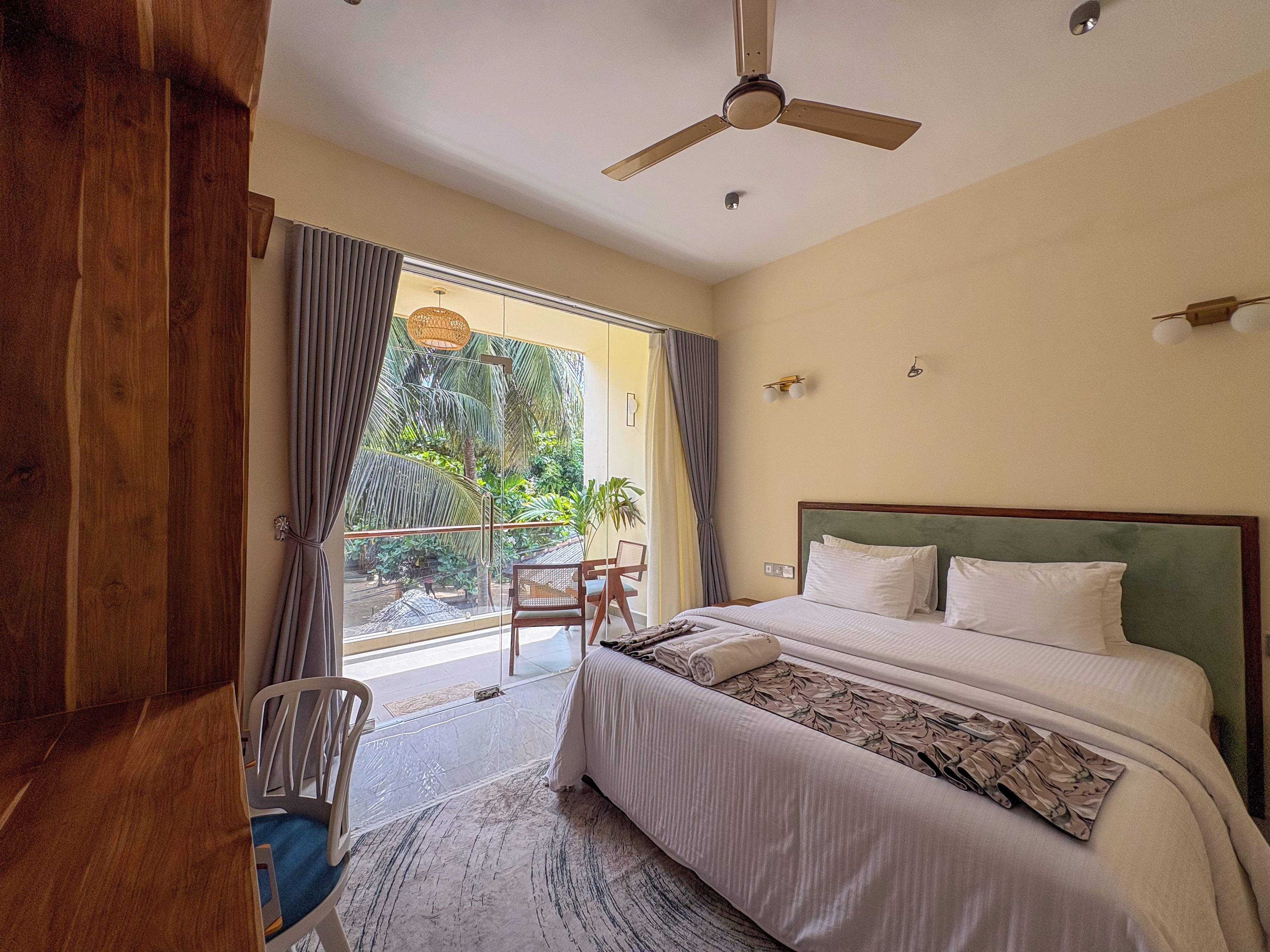Quarto de hotel com uma cama grande, um ventilador de teto, vista para um jardim tropical e uma varanda com uma cadeira.