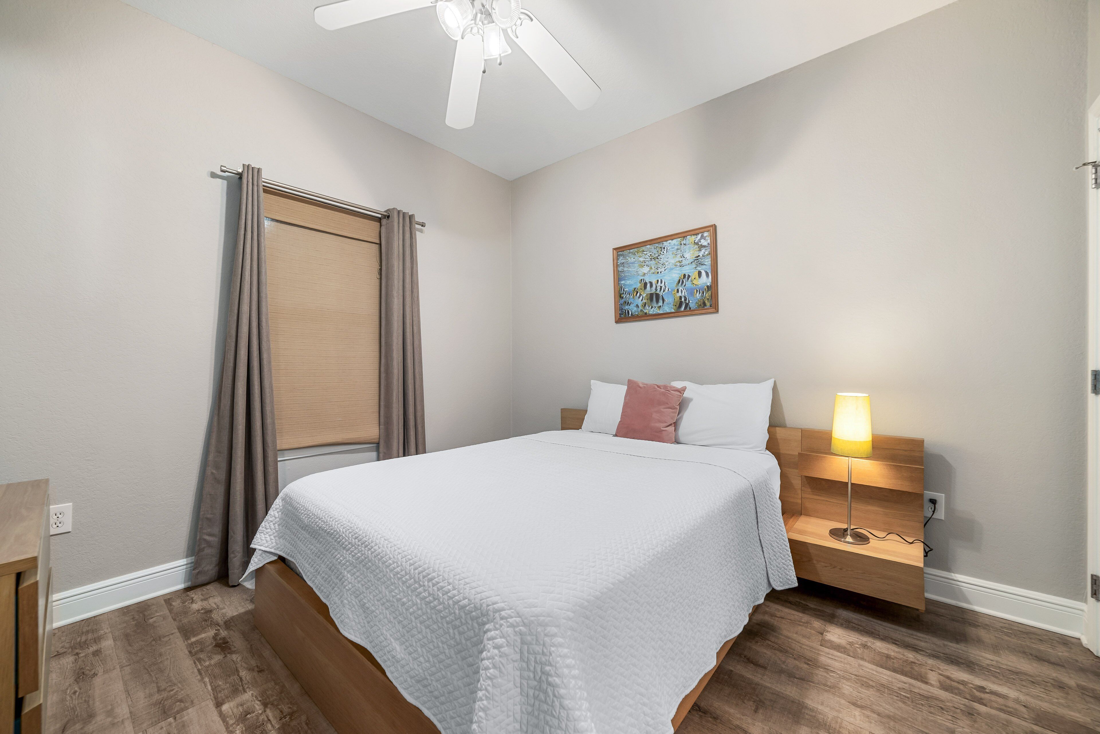 A bedroom with a bed, a nightstand, a lamp, a ceiling fan, a window with curtains, and a painting on the wall.